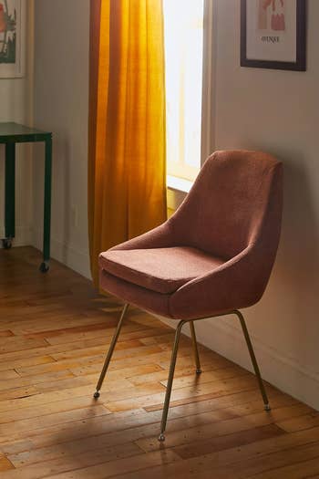 Mid-century modern chair with cushioned seat and metal legs placed in a room with wooden floors and a window covered by a curtain