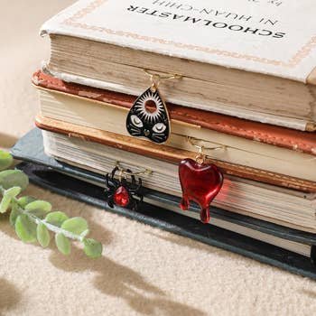 Decorative bookmarks featuring a planchette, a reddish  heart, and a spider with a reddish  gem placed connected  a stack of books