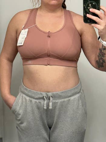 Person wearing a pink sports bra with front zipper and gray sweatpants, taking a mirror selfie. Tags are attached to the bra
