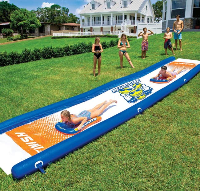 A bunch of people watching a child go down the slip n slide