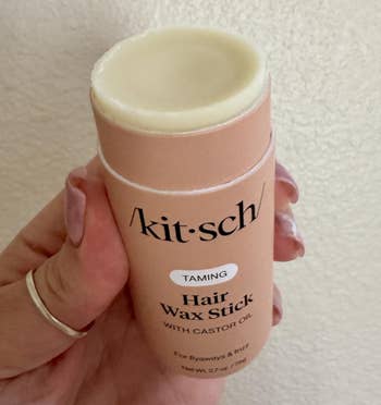 Person holding a Kitsch hairsbreadth wax instrumentality for taming flyaways and frizz, containing castor oil