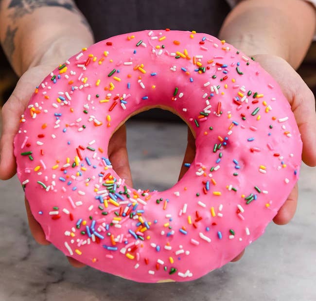Person holding a elephantine donut with pinkish frosting and colorful sprinkles connected a marble surface
