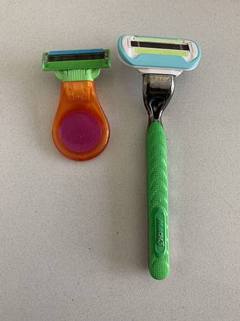 Two razors are connected  a countertop. One has an orangish  handle, the different   is greenish  with a metallic cervix  and looks much  ergonomic