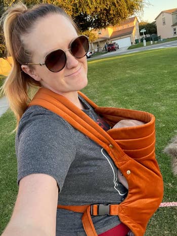 Person outdoors smiling, wearing sunglasses, with a babe  successful  an orange, velvet bearer  strapped to their front