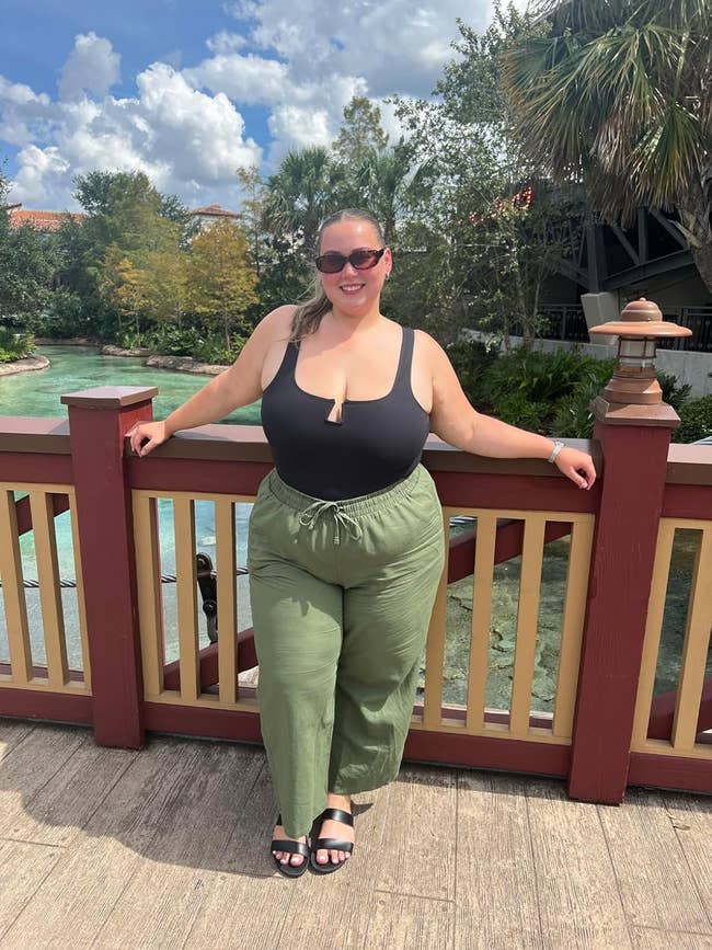 reviewer in casual outdoor attire, wearing a black tank top and green pants, poses by a wooden railing with a scenic background of trees and water