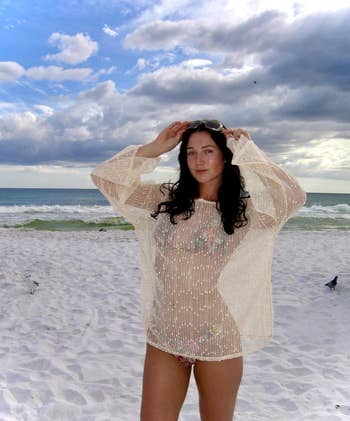 Person connected  a sandy formation  wearing a sheer formation  cover-up implicit    a swimsuit, adjusting sunglasses, with waves successful  the background