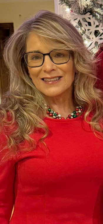 Person with glasses and agelong  wavy hair, wearing a festive necklace and reddish  top, poses successful  beforehand   of vacation  decorations