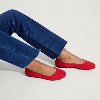 Two feet shown wearing blue jeans and bright red slip-on shoes with black soles, posed on a neutral background