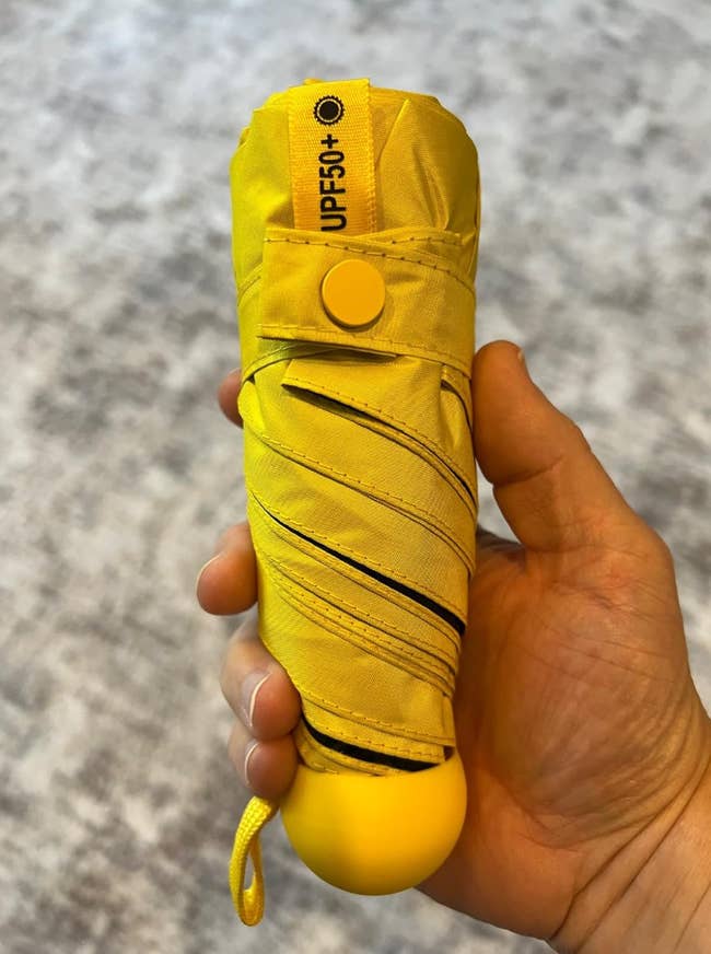 Hand holding a compact, folded yellowish  umbrella with UPF 50+ label, highlighting prima   extortion  diagnostic   for buying  article