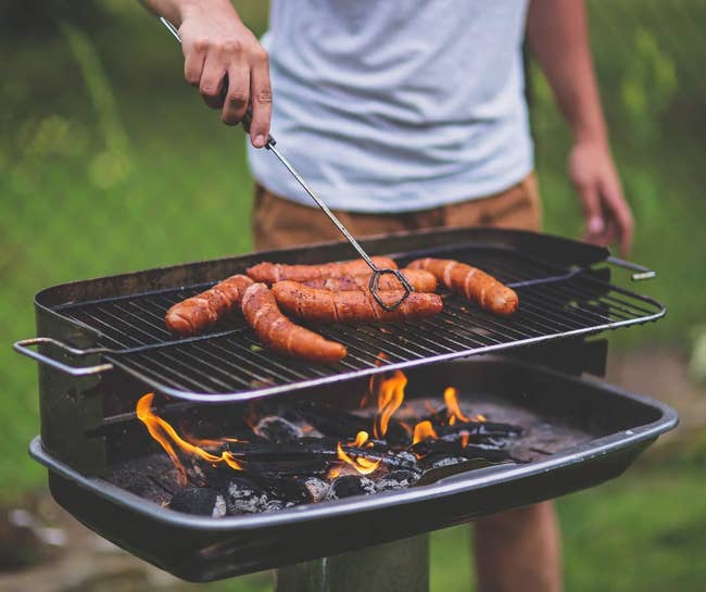 Person grilling sausages on an outdoor barbecue with visible flames, wearing a casual shirt and shorts, in a garden setting