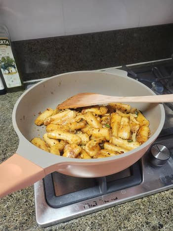 Frying pan with sautéed pasta on a stove, using a wooden spatula; suitable for a kitchenware shopping article