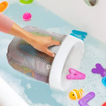 Hand using a gray mesh scoop to collect foam letters and a fish toy in a bathtub