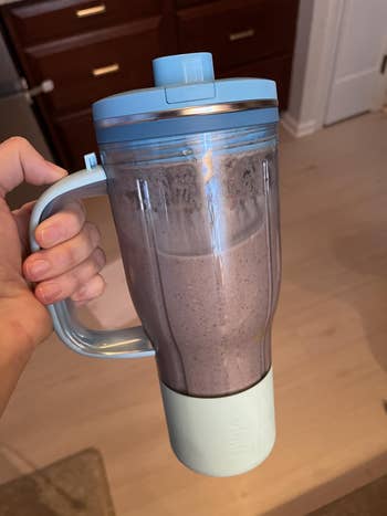 a hand holding the closed tumbler by the handle in the blue raspberry colorway with a blended smoothie inside