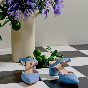 Blue block-heeled shoes with ankle straps connected  a checkered floor, beside a vase of purple flowers and greenish  fruit