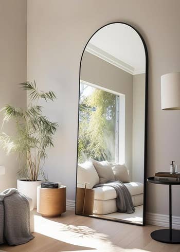Tall arched floor mirror reflecting a room with a cozy nook, plants, and modern furniture, suitable for home decor inspiration