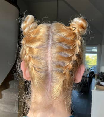 A person with blonde hair braided into two Dutch braids and styled in double buns, showing the back view of their head