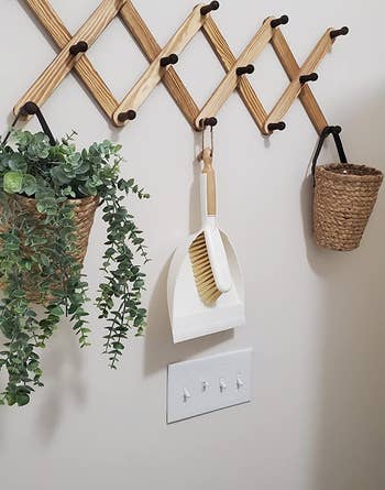 reviewer photo of the white dustpan and brush hanging on a hook next to a plant and a basket