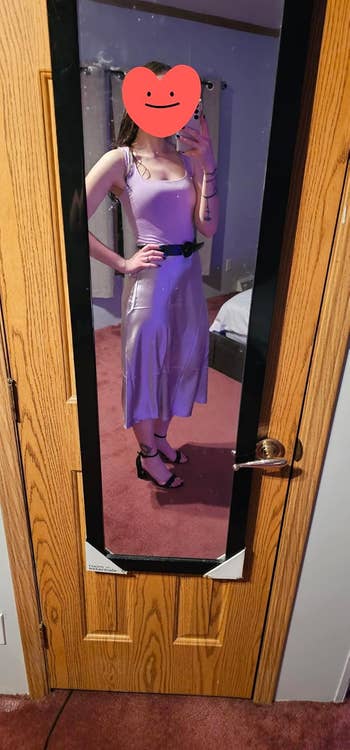 Person taking a mirror selfie, wearing a sleeveless dress with a belt and high heels, standing in a room with a visible bed and door. Face is covered