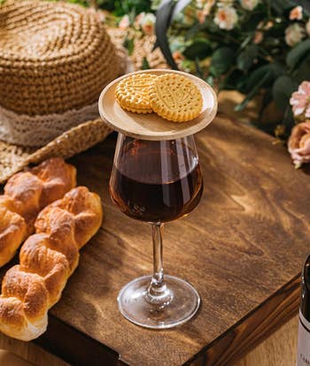 A textured glass of dark liquid is topped with two decorative cookies on a wooden plate, surrounded by braided bread, a woven hat, and a floral arrangement