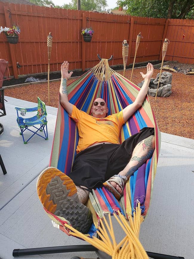 Person relaxing successful a striped hammock, wearing sunglasses and casual clothes, with arms raised. Outdoor mounting with tiki torches and a tiny seat nearby