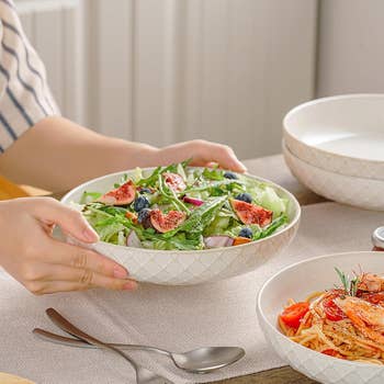 Person serving salad with figs and blueberries in a patterned bowl on a dining table beside another dish