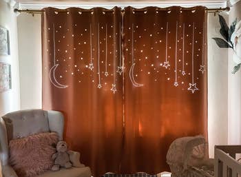 A pink curtain over a window with light streaming through cut outs in the shapes of moons and stars 
