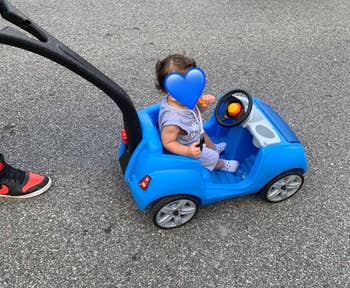 Child in a blue toy car stroller on a sidewalk, accompanied by an adult
