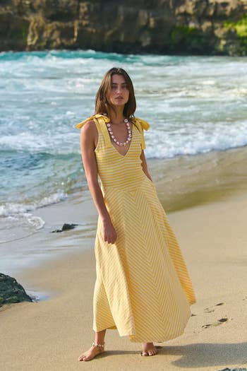 A model on a beach in a sleeveless, long, patterned dress with tie straps, modeling summer fashion