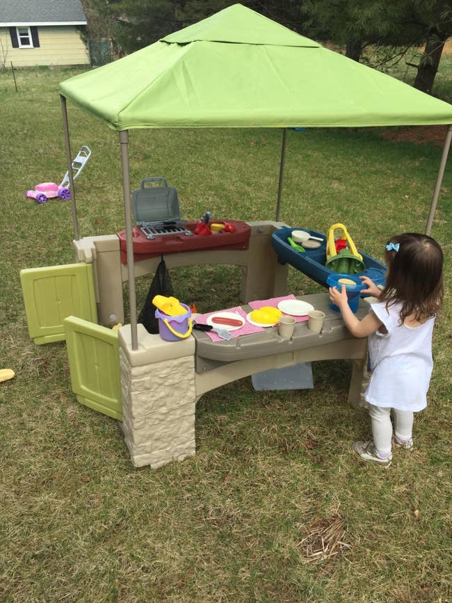 Child playing with a toy kitchen set under a canopy in a backyard