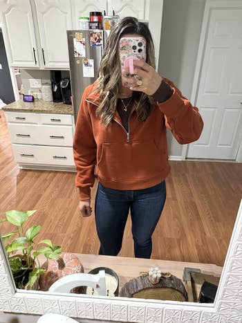 Person taking a mirror selfie in casual outfit: zip-up sweater and jeans, standing in a kitchen. Face obscured by phone