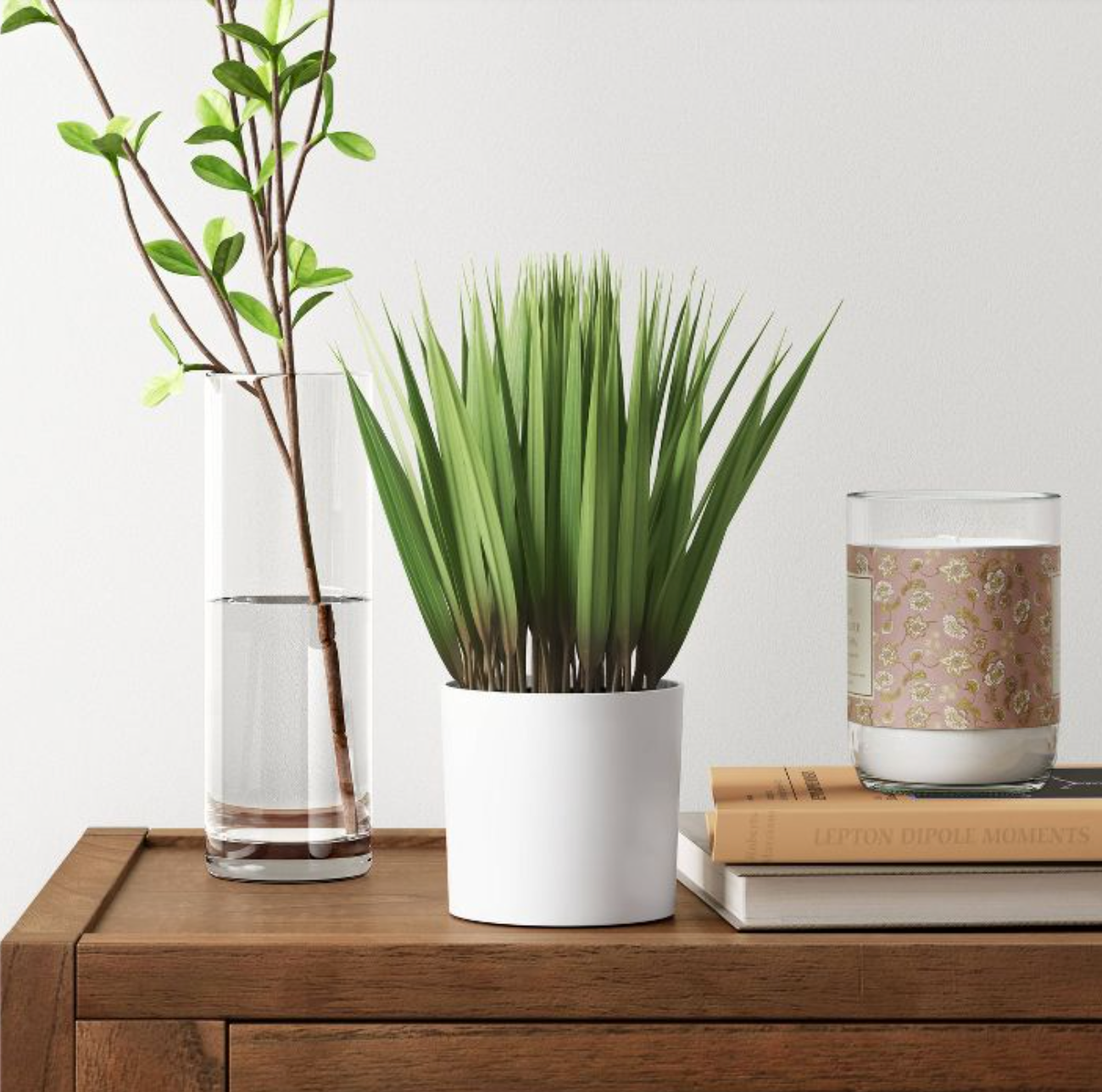 Green fake grass in a white pot on top of a brown cabinet next to a pile of books and a candle