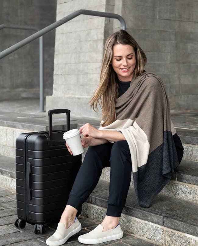A model wearing the brown colorblock scarf while sitting on some stairs with their suitcase