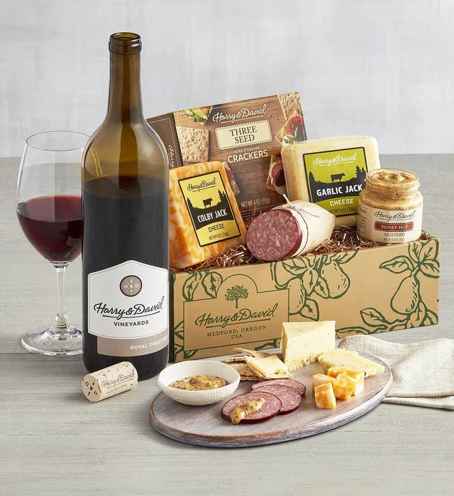 the gift box with cheeses, and a mini charcuterie with meats and crackers with a glass of wine next to the bottle of wine