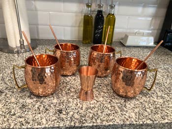 Four copper Moscow mule mugs with straws and a matching jigger on a granite countertop, with kitchen items in the background