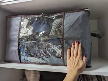 A hand rests on a clear, zippered storage organizer with folded clothes visible inside, placed on a shelf