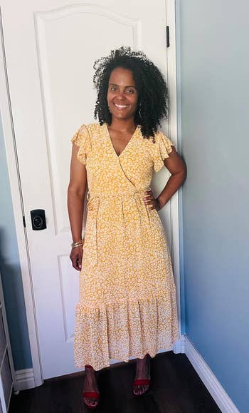 revi smiling in a light, floral wrap dress with ruffled sleeves and hem, standing in a room with a door in the background