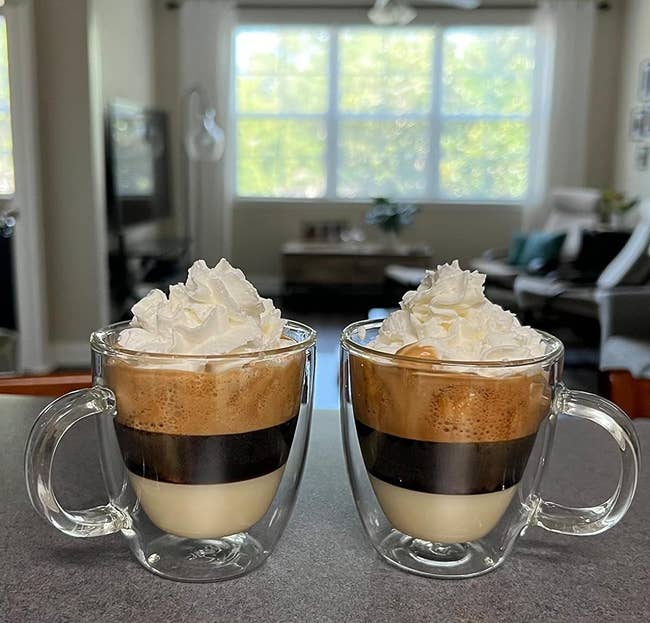 A reviewer photo of two layered coffee drinks topped with whipped cream in clear mugs on a kitchen counter