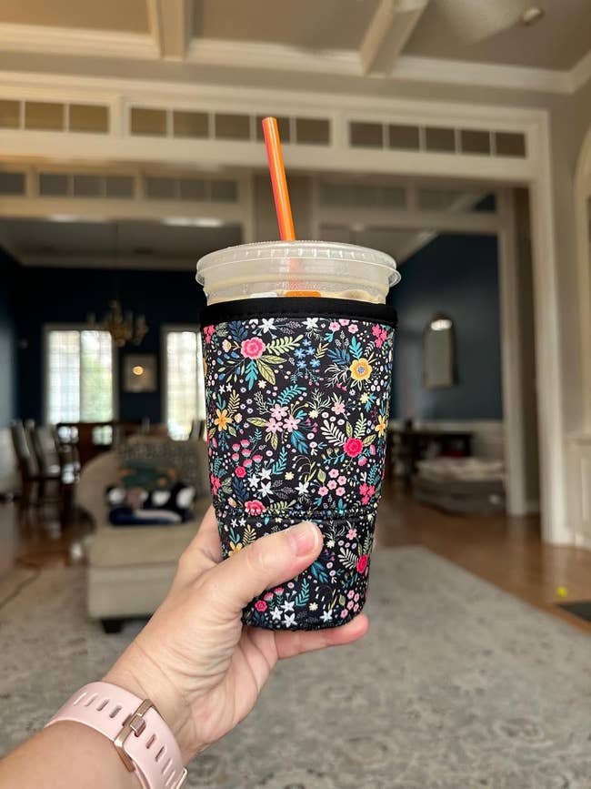 Hand holding iced drink in floral-patterned sleeve with straw, inside a spacious living room