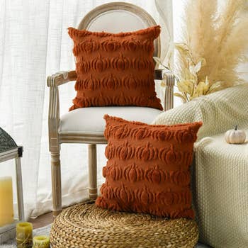 Two textured pillows with a pumpkin motif displayed on a chair and basket in a cozy room setting, enhancing fall-themed home decor