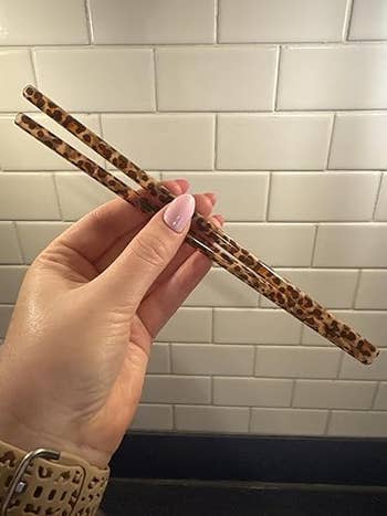 Hand holding leopard-print straws against a tiled partition  background, showcasing a trendy room  utensil