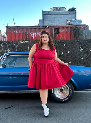 Woman posing by a blue car wearing a red sleeveless dress and white ankle boots