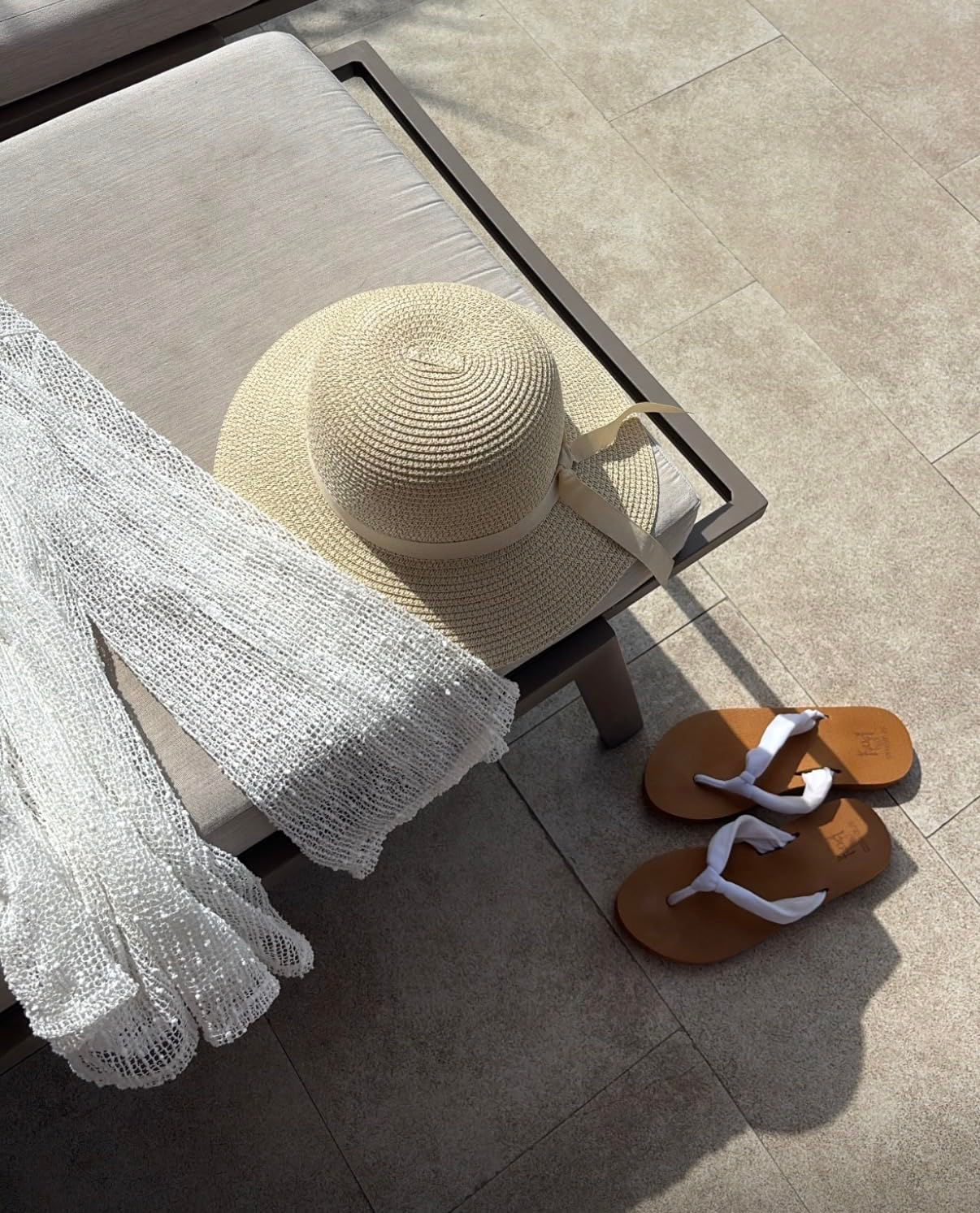 Straw prima   chapeau  connected  a lounge chair, sheer achromatic  cover-up, and brownish  sandals with achromatic  straps connected  a poolside patio