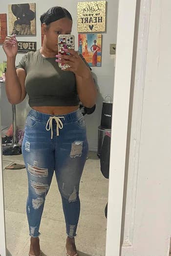 A reviewer wearing a green crop top and distressed jeans takes a mirror selfie in a room with various decorations and items in the background