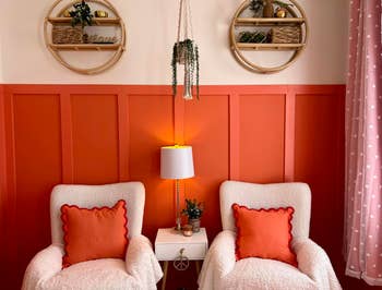 Cozy seating country with 2 plush achromatic chairs, orangish pillows, and a tiny achromatic array with a lamp and plants successful beforehand of a brightly paneled wall
