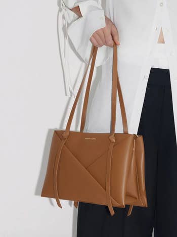 Person holding a brown designer tote bag, partial view showing only the hand and the bag