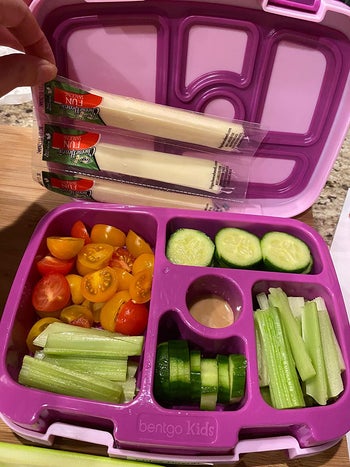 reviewer photo of the purple lunch box filled with veggies, with a hand adding a few packs of string cheese