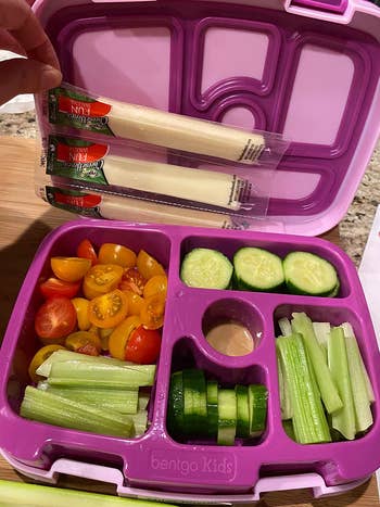 reviewer photo of the purple lunch box filled with veggies, with a hand adding a few packs of string cheese