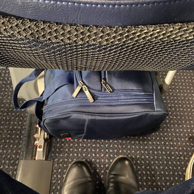 A blue bag placed under an airplane seat, with a person wearing black shoes