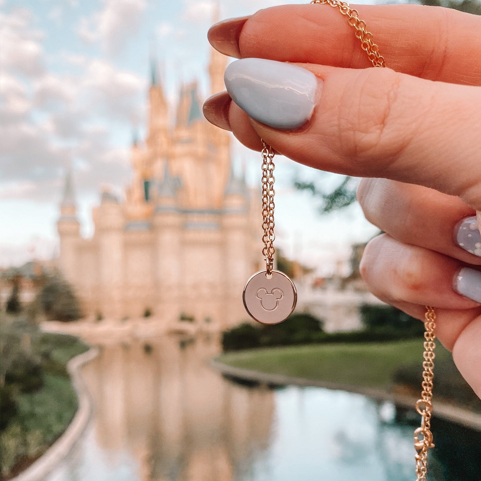 a dainty circle charm with an outline of mickey in the middle being held in front of the castle