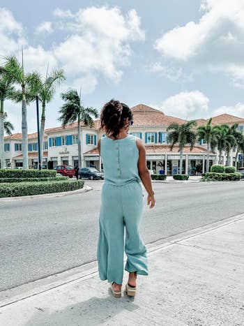 Person walking on a sunny street, wearing a sleeveless jumpsuit and sandals, with a palm tree-lined shopping center in the background